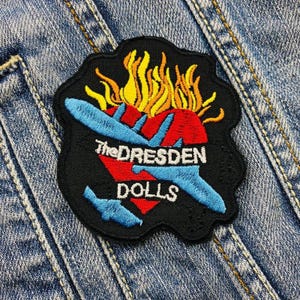 May include: Black and red embroidered patch with a blue airplane and flames. The text "The Dresden Dolls" is written in white on the patch.
