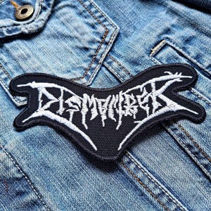 Könnte beinhalten: Schwarz-weiß gestickter Patch mit dem Wort "DEMOLISHER" in einer stilisierten Schriftart. Der Patch ist auf eine blaue Jeansjacke genäht.