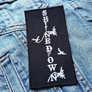 Op de afbeelding: Zwarte patch met witte geborduurde tekst die "Shine Down" luidt met gestileerde bloemmotieven en vogels aan weerszijden van de tekst.