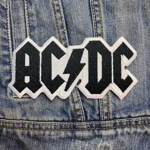 Könnte beinhalten: Hellblaue Jeansjacke mit einem großen, gestickten Aufnäher. Der Aufnäher zeigt den Bandnamen "AC/DC" in fetten, schwarzen Buchstaben mit einem weißen Blitz. Die Jacke hat sichtbare Nähte.