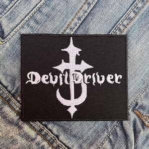 May include: A black rectangular patch with the words "Devil Driver" in white gothic lettering. The patch features a white cross-like design with a crescent moon shape at the bottom. The patch is on a denim background.
