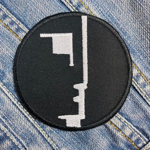 May include: A round, black embroidered patch featuring a white abstract design. The design includes a vertical line and a flag-like shape. The patch is set against a denim background.