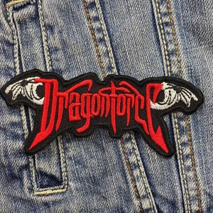 Może przedstawiać: Naszywka haftowana w kolorze czarnym i czerwonym z napisem "Dragonforce" w czerwonych literach i dwoma białymi skrzydłami smoka po obu stronach.