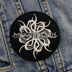 Op de afbeelding: Een ronde, zwarte patch met het woord "MESHUGGAH" in witte, gestileerde letters. De patch heeft een wit, abstract ontwerp boven de tekst, dat lijkt op tentakels. De patch is geplaatst tegen een denim achtergrond.