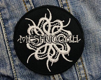 Może przedstawiać: Okrągła, czarna naszywka z białym napisem "MESHUGGAH" w stylizowanej czcionce. Naszywka posiada biały, abstrakcyjny wzór nad tekstem, przypominający macki. Tło z dżinsu.