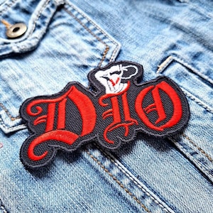 Op de afbeelding: Een zwart en rood geborduurd patch met het woord "DIO" in een gestileerd lettertype. Een kleine witte hand met een vuist bevindt zich boven de "D".