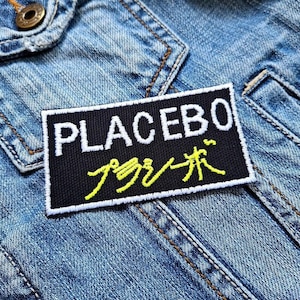 May include: A black patch with white text that reads "PLACEBO" and yellow Japanese text. The patch is sewn onto a blue denim jacket.