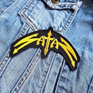 May include: A black and yellow embroidered patch with the word "SATAN" in a stylised font.