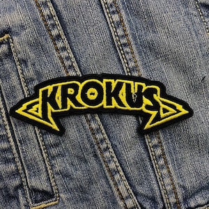 Könnte beinhalten: Schwarzer und gelber gestickter Aufnäher mit dem Wort "KROKUS" in einer stilisierten Schrift.