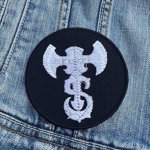 May include: White embroidered axe design on a black circular patch. The patch is made of fabric and has a stitched edge.