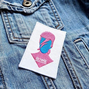 Peut inclure: Écusson brodé avec une silhouette rose et bleue d'une personne, le mot "BOWIE" en blanc. L'écusson est rectangulaire avec un fond blanc, placé sur une veste en jean.