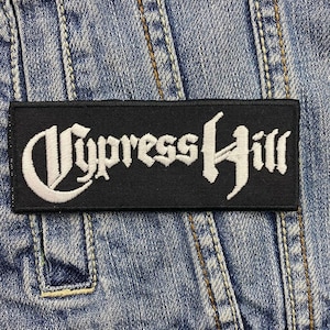 Op de afbeelding: Een rechthoekige zwarte patch met de tekst "Cypress Hill" in witte, gestileerde letters. De patch is genaaid op een spijkerjasje, met zichtbare stiksels en een zakdetail.