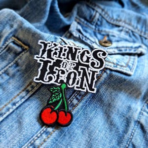 May include: Black and white embroidered patch with the text "Kings of Iron" and a green stem with two red cherries.