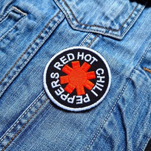 Puede incluir: Un parche circular con la inscripción "RED HOT CHILI PEPPERS" en blanco sobre fondo negro y un diseño de asterisco naranja. El parche está cosido a una chaqueta vaquera azul.