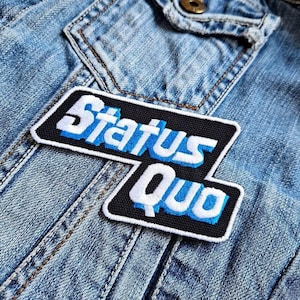 Könnte beinhalten: Ein gestickter schwarzer und weißer Aufnäher mit dem Text "Status Quo" in blauen und weißen Buchstaben. Der Aufnäher ist auf eine blaue Jeansjacke genäht.