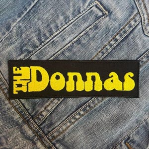 May include: A black rectangular patch featuring the yellow text "THE Donnas". The patch is made of woven fabric and is set against a denim background. The text is in a bold, stylised font.