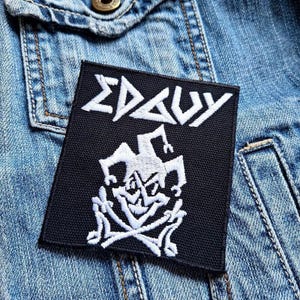 Op de afbeelding: Zwart-witte geborduurde patch met de tekst "EDGUY" en een wit narrenontwerp. De patch is genaaid op een blauw denim jack.