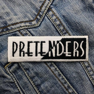 Puede incluir: Parche bordado blanco y negro con la palabra "PRETENDERS" en letras negras sobre fondo blanco.