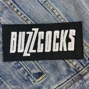 May include: Black and white embroidered patch with the text "Buzzcocks" in white lettering.