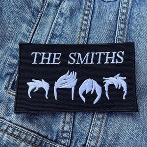 Peut inclure: Patch brodé noir et blanc avec le texte "THE SMITHS" en lettres blanches. Le patch présente quatre silhouettes de cheveux blanches stylisées.