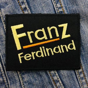 May include: A black rectangular patch with the name "Franz Ferdinand" embroidered in yellow thread. The word "Franz" is above "Ferdinand", with an orange line separating the two words. The patch is set against a denim background.