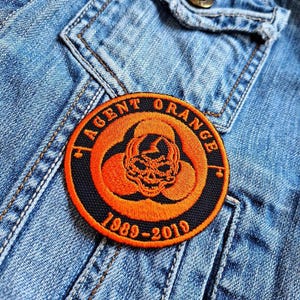 May include: A black and orange embroidered patch with the text "Agent Orange 1989-2010" and a skull design.