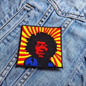 Könnte beinhalten: Ein gestickter Aufnäher in Schwarz und Rot mit einem stilisierten Bild einer Person mit einem großen Afro und einem roten Gesicht. Der Hintergrund ist ein gelbes und rotes Sonnenstrahlenmuster.