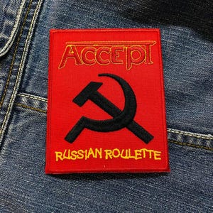 Può includere: Toppa ricamata rossa con il nome della band "ACCEPT" in lettere gialle e le parole "RUSSIAN ROULETTE" in lettere gialle sotto un'immagine in bianco e nero di un martello e una falce.