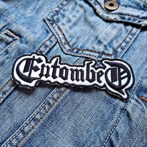 May include: A black and white embroidered patch with the word "Entombed" in a gothic font. The patch is on a blue denim jacket. The patch has a white border and is rectangular in shape.