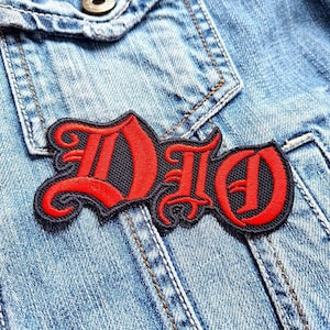 May include: Red and black embroidered patch with the word "DIO" in a gothic font style. The patch is sewn onto a blue denim jacket.
