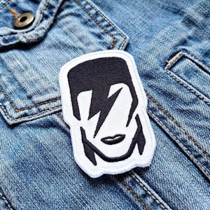 May include: Black and white embroidered iron-on patch of a stylized face with a lightning bolt across the eye. The patch is on a blue denim jacket.