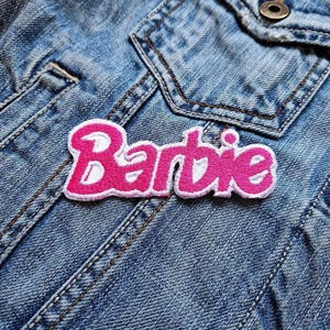 以下が含まれることがあります： 青いデニムジャケットにピンクの「Barbie」アイロン接着パッチ