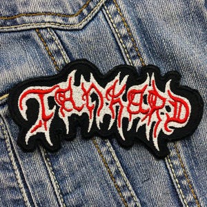 May include: A black embroidered patch with the word "TANKARD" in red and white, featuring a stylized, jagged font. The patch is sewn onto a blue denim jacket.