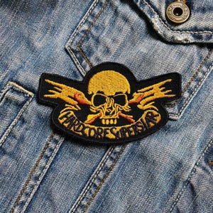May include: Embroidered patch with a yellow skull and orange details, and the words "HARDCORE SUPERSTARS" on a black base. The patch is attached to a denim jacket.