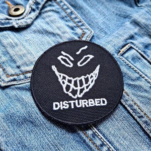 Könnte beinhalten: Schwarz-weiß gestickter Patch mit einem grinsenden Gesicht und dem Wort "DISTURBED" darunter. Der Patch befindet sich auf einer blauen Jeansjacke.