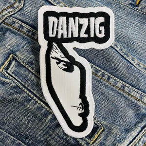 Op de afbeelding: Een wit en zwart geborduurde patch met het woord "DANZIG" boven een gestileerd gezicht. De patch heeft een zwarte rand en is geplaatst tegen een denim achtergrond.