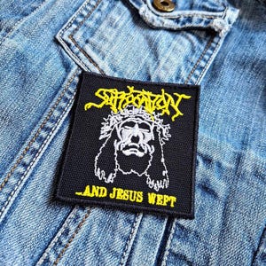 Op de afbeelding: Een zwarte vierkante patch met de gele tekst "Sufferation" boven een witte illustratie van een figuur met een doornenkroon. De tekst "...AND JESUS WEPT" staat eronder. De patch bevindt zich op een blauw spijkerjasje.