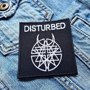 May include: A black square patch featuring the word "DISTURBED" in white capital letters. Below the text is a white geometric emblem. The patch is displayed on a blue denim jacket.