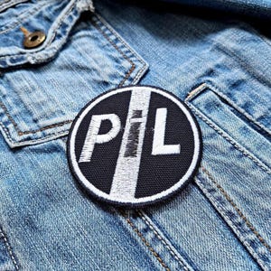 May include: A round, black and white embroidered patch with the letters "P" "I" and "L" in white. The patch is on a denim jacket. The letters are in a bold, sans-serif font. The patch has a white border.