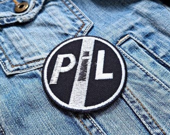 Può includere: Una toppa rotonda ricamata in bianco e nero con le lettere "P", "I" e "L" in bianco. La toppa è su una giacca di jeans. Le lettere sono in grassetto e hanno un bordo bianco.