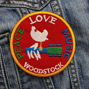 Op de afbeelding: Een geborduurde patch met de woorden "Peace Love Music Woodstock" en een witte duif die een gitaar vasthoudt, in rood, geel en groen.