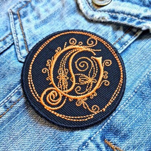 May include: A round, navy blue embroidered patch with an ornate, orange letter "O" in the center. The letter is surrounded by decorative swirls and floral elements. The patch is set against a denim jacket.