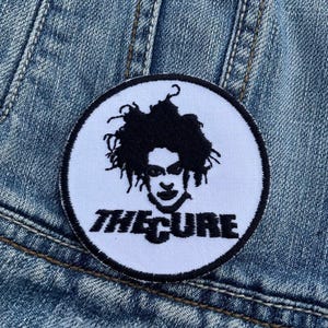 May include: White embroidered patch with a black and white image of Robert Smith, lead singer of The Cure, and the band's name "The Cure" in black lettering.