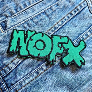 May include: A green embroidered patch with the word "NOFX" in a stylized font, outlined in black. The patch is set against a backdrop of blue denim jeans, showcasing the texture of the fabric and stitching.