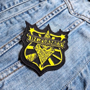 May include: Black and yellow embroidered patch with the word "IN FLAMES" in a banner across the top. The patch is shaped like a shield with a star at the top and bottom.