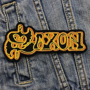 Puede incluir: Parche bordado negro y amarillo con la palabra "Saxon" en una fuente estilizada.