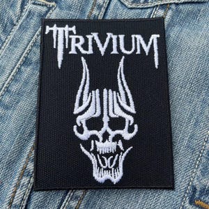 Könnte beinhalten: Schwarzer rechteckiger Aufnäher mit dem Wort "TRIVIUM" in Weiß, über einem weißen Totenkopf-Design. Der Aufnäher befindet sich auf einem blauen Jeans-Hintergrund. Der Aufnäher ist wahrscheinlich für eine Jacke oder eine Tasche.