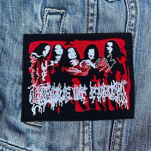 May include: Black rectangular patch with a red and white design. The patch features a stylized illustration of figures and the text "CRADLE OF FILTH" in white, gothic-style lettering. The patch is set against a denim jacket.