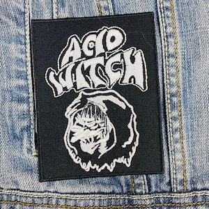 May include: Black and white patch with the text "Acid Witch" in a stylized font. The patch features a hooded figure with a skull-like face.