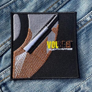 May include: Square embroidered patch with a black background and a stylized design. The design features a white and black element, a brown shape, and the word "VOLBEAT" in yellow. The patch has a stitched border.